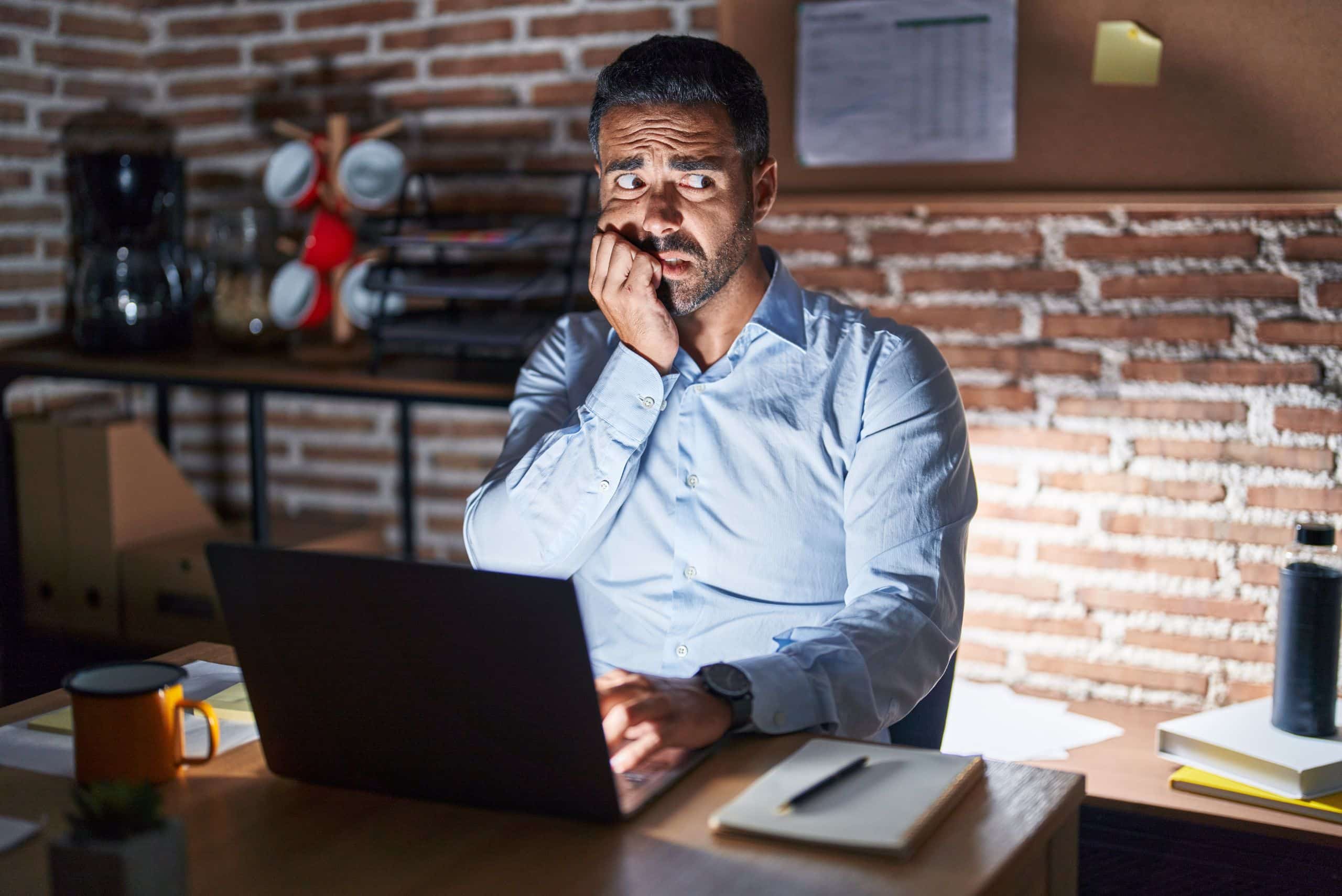an with a laptop sitting at a desk in a dark room looking scared